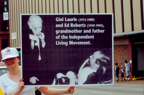 A woman holds up a sign that depicts Gini Laurie and Ed Roberts and includes the caption 'Gini Laurie and Ed Roberts, grandmother and father of the Independent Living Movement.'