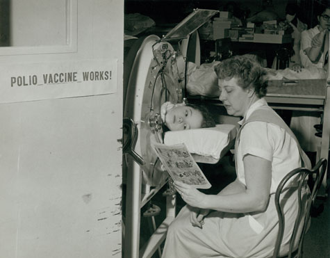 A child in an iron lung is read to by a nurse.