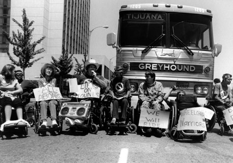 A group of people in wheelchairs gathers in front of a Greyhound bus with signs that read 'All Aboard,' 'We will ride,' and 'Lifts on Hounds.'