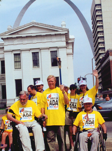 Max Starkloff, Representative Richard Gephardt, Colleen Kelly Starkloff, and Jim Tuscher stand together downtown in the shadow of the St. Louis Arch. Gephardt holds aloft the torch from Spirit of the ADA Torch Relay.